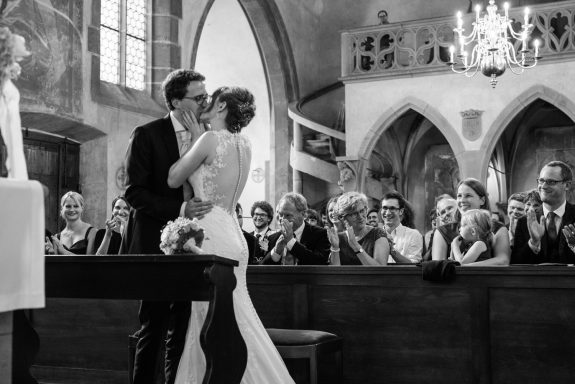 Brautpaar in einer historischen Kirche küsst sich vor den applaudierenden Gästen.