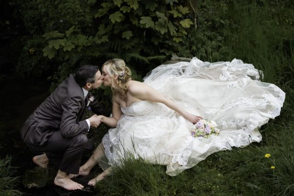 Brautpaarshooting auf einer Wiese. Die Braut trägt ein weites Hochzeitskleid mit Spitze. Beide sind barfuß und sitzen im Graß.